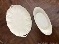 Two white cream colored ceramic dinnerware pieces on brown wooden table: a 17 inch Lenox platter shaped like a scallop shell with gold trim and cutout handles; a 14 inch oval bread plate with a basket weave texture rim.