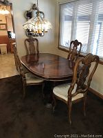 Dining table and four chairs arranged around the table under a stained glass style chandelier. Table is polished wood with an extendable leaf inserted.