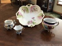 Photo shows the large decorative bowl with floral design, mustache cup, small decorative cup, and medium large ornate cup on a wooden surface in ambient indoor light.