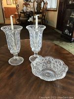 Two clear glass candle holders with intricate vertical cut glass patterns, each holding a white taper candle, placed on a wooden table with decorative clear glass bowl beside them.