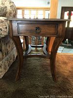 Side view of solid wood side table showing curved legs, stretcher, and drawer with metal ring pull handle.