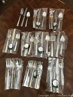 Full view of multiple flatware packages laid out on a round table showing forks, knives, spoons in sealed plastic wrapping including extra spoons and serving pieces