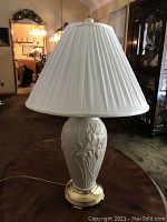 Full view of Lenox porcelain lamp with raised floral designs, brass-tone base, and white pleated lamp shade.