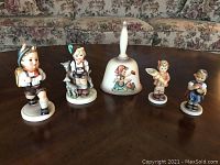 Front view of five Hummel Goebel items on a wooden table including four figurines and a ceramic bell.