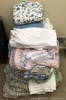 Stack of assorted linens including floral, striped sheets, white sheets, and a pastel colored handmade afghan wrapped in plastic