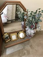 Large wooden framed wall mirror with three small gold-tone framed mirrors on the glass shelf beneath it and floral vase arrangement to the side.
