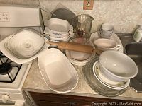 Overview of assorted kitchenware including Corningware dishes stacked with lids, Pfaltzgraff serving dishes, a glass pitcher, rolling pin, and pie plates arranged on kitchen counter near stove and sink.