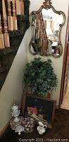 Photo of stairway corner showing gold-tone ornate wall mirror, artificial ivy plant, tall white urn with lid, and decorative boxes and ceramic pitcher in front of wall below stair rail.