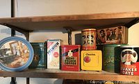 Wide shot of multiple assorted vintage tins lined up on wooden shelf showing varied designs and brands.