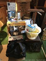 Wide view showing Mikasa crystal glassware set, Presto electric griddle box, sandwich maker box, brass candlestick, gold trays, Tupperware basket, and black bags.