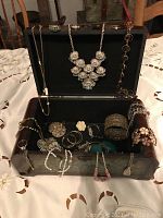 Photo of open wooden jewelry box with black felt interior showing assorted jewelry including large silver-tone floral necklace, green stone bangles, bracelets, earrings, and brooches.