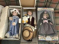 Three boxed dolls displayed in a row on a plaid couch. From left to right: a doll in blue dress with bonnet, a male doll holding a white duck plush and a straw hat, and a boxed Victorian Collection doll in black and pink polka dot dress with bonnet.