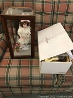 Porcelain doll inside wooden glass display case with accompanying box on couch