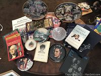 Image shows an assortment of decorative plates laid out on a round table along with several books and printed material. Plates feature historical and celebrity figures including presidents, Elvis Presley, Diana, and religious subjects. A large crystal covered dish is also visible.
