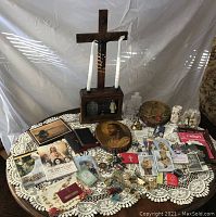 Full view of entire religious lot on lace tablecloth showing wooden cross with candle holders and many cards, booklets, plaques, rosaries, and figurines arranged around it.