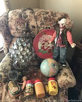 Full view of vintage assortment items arranged on floral patterned armchair showing hanging lamp, clown doll, globe, thermoses, and Strawberry Shortcake platter.