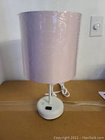 Front view of stick lamp with purple lampshade wrapped in plastic and white base on cardboard surface