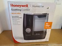 Front view of original boxed Honeywell warm mist humidifier showing branding, key features like bacteria free mist up to 99.5%, no filter required, auto shut off, wide tank opening, and essential oil feature.