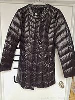 Front view of the dark asymmetrical puffer jacket on a hanger, showing shiny quilted fabric with diagonal lines and snap button closure.