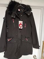 Full view of black long jacket with faux fur hood hung on hanger showing front with zipper and button placket, zipper pockets and drawstring waist.