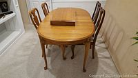 Photo showing solid oak dining table with two leaves on top and four dining chairs, two on each side of the table. The table is oval with ornate legs.