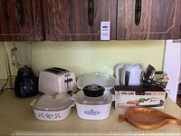 Photo shows multiple kitchen appliances arranged on a counter, including Black & Decker Crushmaster blender, Proctor Silex toaster, electric carving knife, Sunbeam kettle, GE steam iron, casserole dishes, and knife set in box