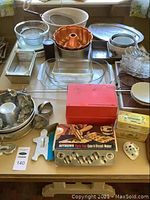 Photo shows a collection of baking pans and kitchen tools including glass mixing bowls, metal cake pans, cookie cutters, baking dishes, a warming tray, and boxes containing cake and biscuit maker and cookbooks.