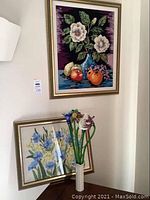 Overview of two framed needlepoint artworks and glass floral arrangement in ceramic vase on floor and leaning against wall