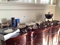 Photo showing an array of silver plated serve ware on a wooden sideboard, including chafing dish, covered serving dishes, cream and sugar set, trays, and plates.