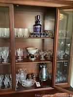 Wooden cabinet holding an assortment of crystal glassware including decanter, shot glasses, coffee carafe, stemmed glasses, tumblers, and bowls.