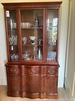 Front view of entire dining hutch showing glass top with three shelves inside and wooden base with drawers and cupboard doors.