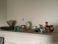 Photo showing a variety of vintage decorative glass and porcelain items on a mantel, including vases and pitchers and a clock in background.
