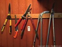 Overview of all five yard tools hanging on a wall showing knives, clippers, saw, and loppers.