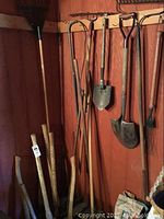Wide angle photo showing various yard tools hanging on wall including rakes, shovels, hoe, and folding shovel along with wooden handled tools stored upright in a corner.