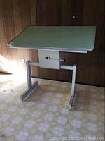 Full view of Radius Tension drafting table angled with pale green tabletop and white metal base on patterned linoleum floor against wood panel wall.