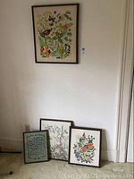 Four framed pieces including three needlepoint artworks and one beveled mirror, arranged with one hanging and three leaning against wall