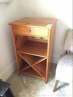 Front view of wooden bar cabinet showing single drawer, wine glass rack beneath it, open shelf and X-shaped wine bottle storage at bottom.