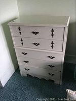 Front view of white 5-drawer dresser showing decorative dark metal handles and scalloped apron base.