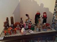 Multiple hand painted and decorative figurines arranged on a wooden dresser with lace covering. Includes various cultural dolls and figures.