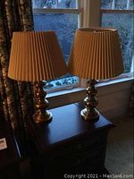 Pair of brass table lamps with pleated beige fabric shades on a wooden surface near a window. Both lamps are 26 inches tall. One shade has a small hole, visible on closer inspection.