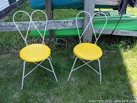 Photo of two white wrought iron chairs with yellow vinyl round seats placed on grass outdoors with a wooden fence and green kayak in background.