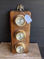 Full front view of vintage weather station showing wooden plaque with brass eagle and three brass circular dials vertically mounted.