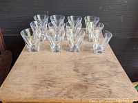 Set of 12 clear heavy glass tumblers with round rims and square bottoms arranged on a wooden surface.