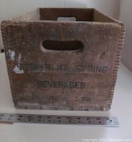 Front view of wooden crate with 'Hosmer Mt. Spring Beverages' stencil, showing cutout hand grip and ruler for scale.
