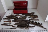 Red metal toolbox open showing a variety of large metal auger drill bits laid out in front.
