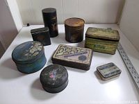 Overview of all 8 metal tins and boxes, assorted shapes and sizes, showing decorative labels and painted designs, surface wear and patina visible