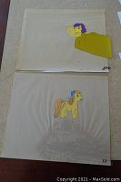 Two animation cells on table; one of yellow pony with purple mane under a yellow blanket, other of yellow pony with orange mane and blue ribbon