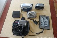 Photo showing all electronic devices included in the lot on a beige surface: Kindle, two digital camcorders, GPS device with headphones and case, Apple TV box, and a charging cable.