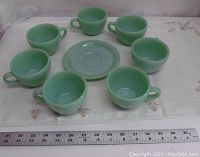 Seven mint green Fire King ovenware tea cups arranged around one matching saucer on a white cloth with ruler for size reference.