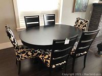 Six chairs arranged around a black round table on dark wood floor near window